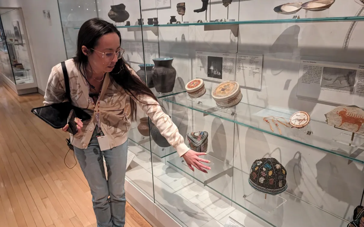 An Indigenous woman with long, dark hair and glasses stands by a museum case filled with belongings showing beadwork, quillwork, and other artistic traditions.
