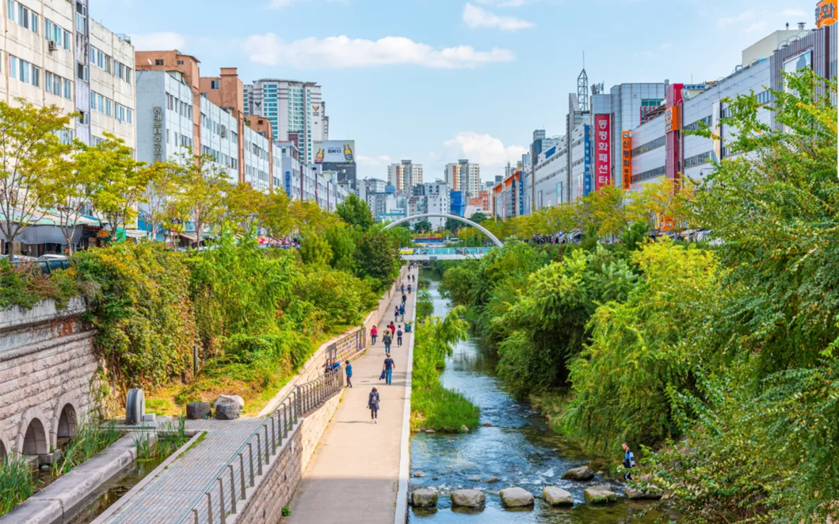 Urban park with a walking path, railway tracks on the left, trees, and a river flowing through the middle under a blue sky.