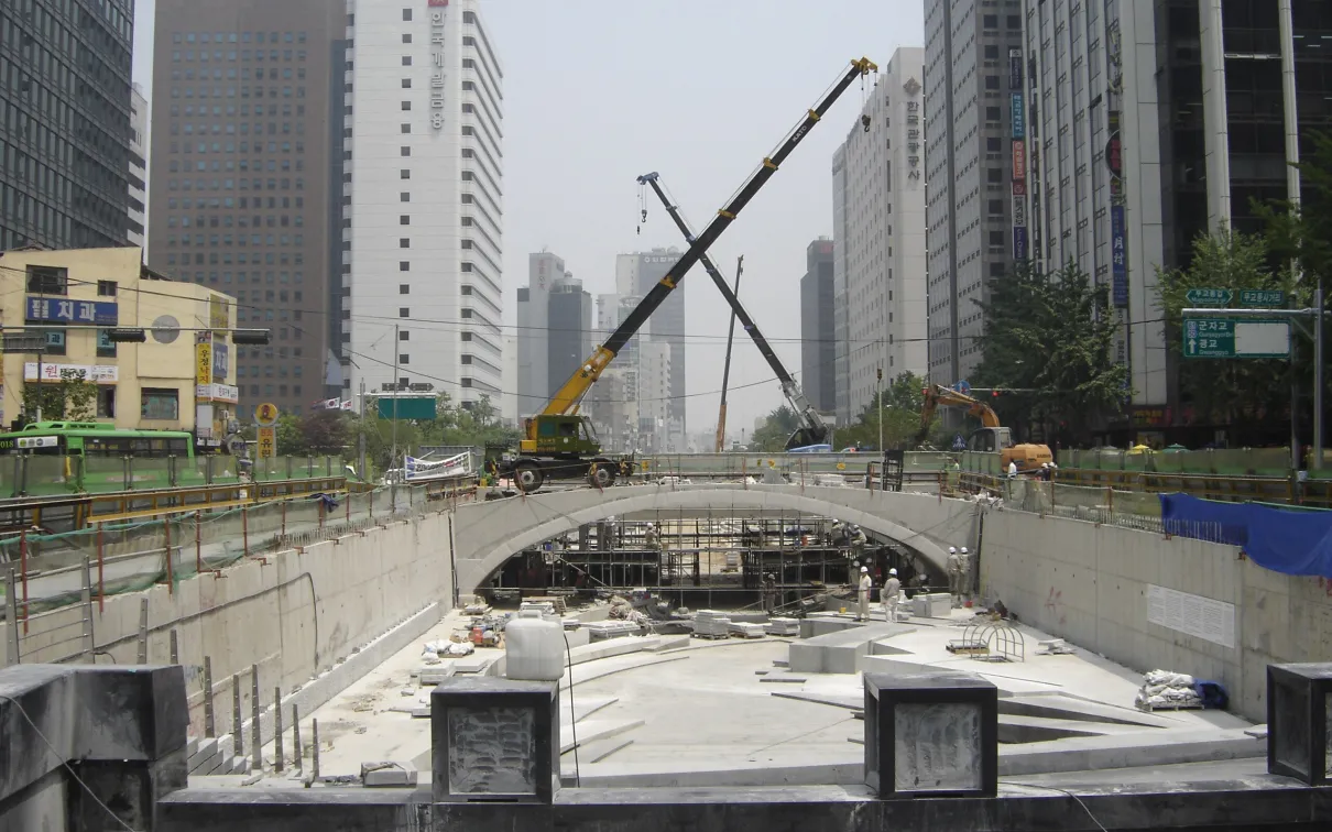 Construction site of an underground structure between tall buildings with cranes and rebar visible.