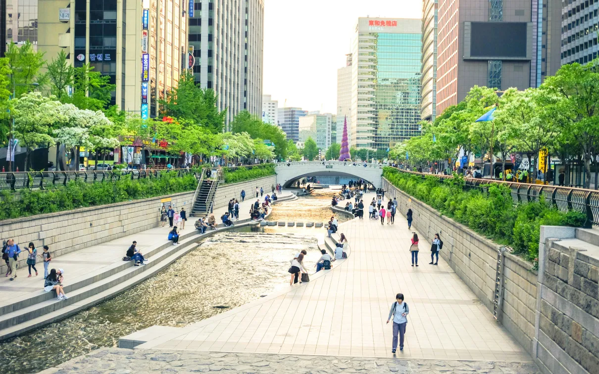 Urban waterway with stone steps on both sides, people walking and sitting along the stream in a city setting.