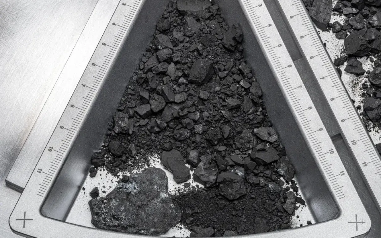 Pile of black coal-like chunks inside a white triangular container marked with a "+" sign.