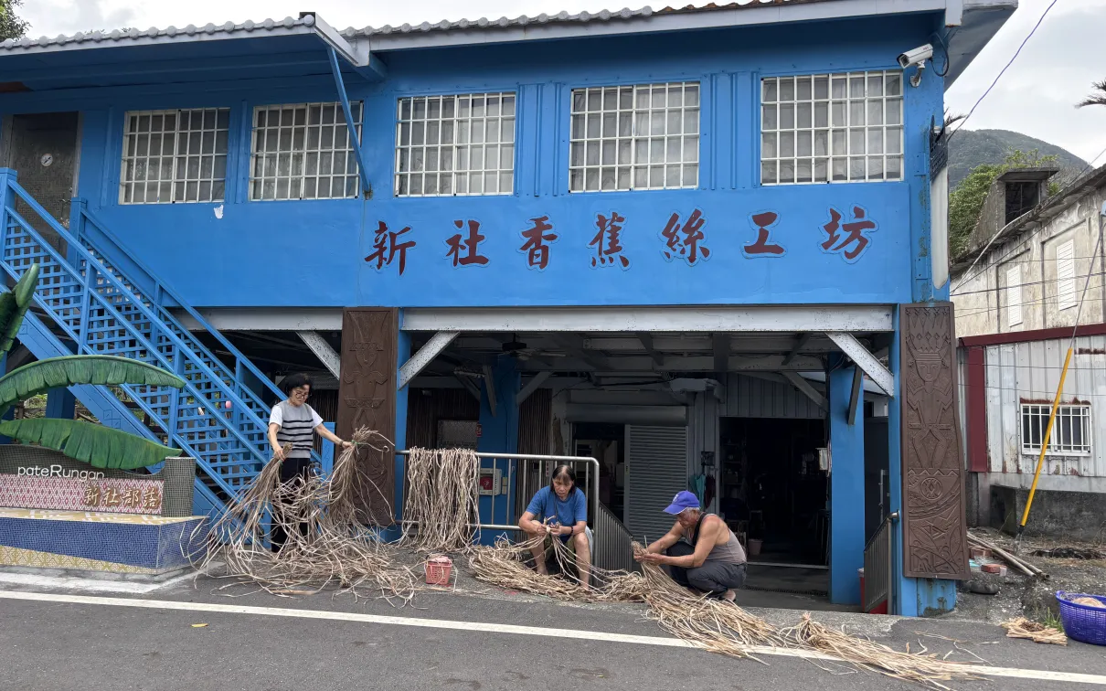 Three people sit outside a blue building sorting and weaving bundles of dried plant fibers along the roadside.