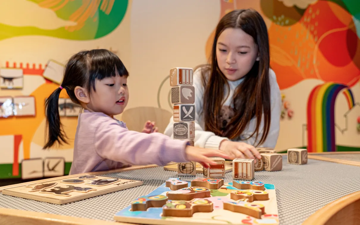 Two children playing with wooden blocks at a table inside a colorful room.
