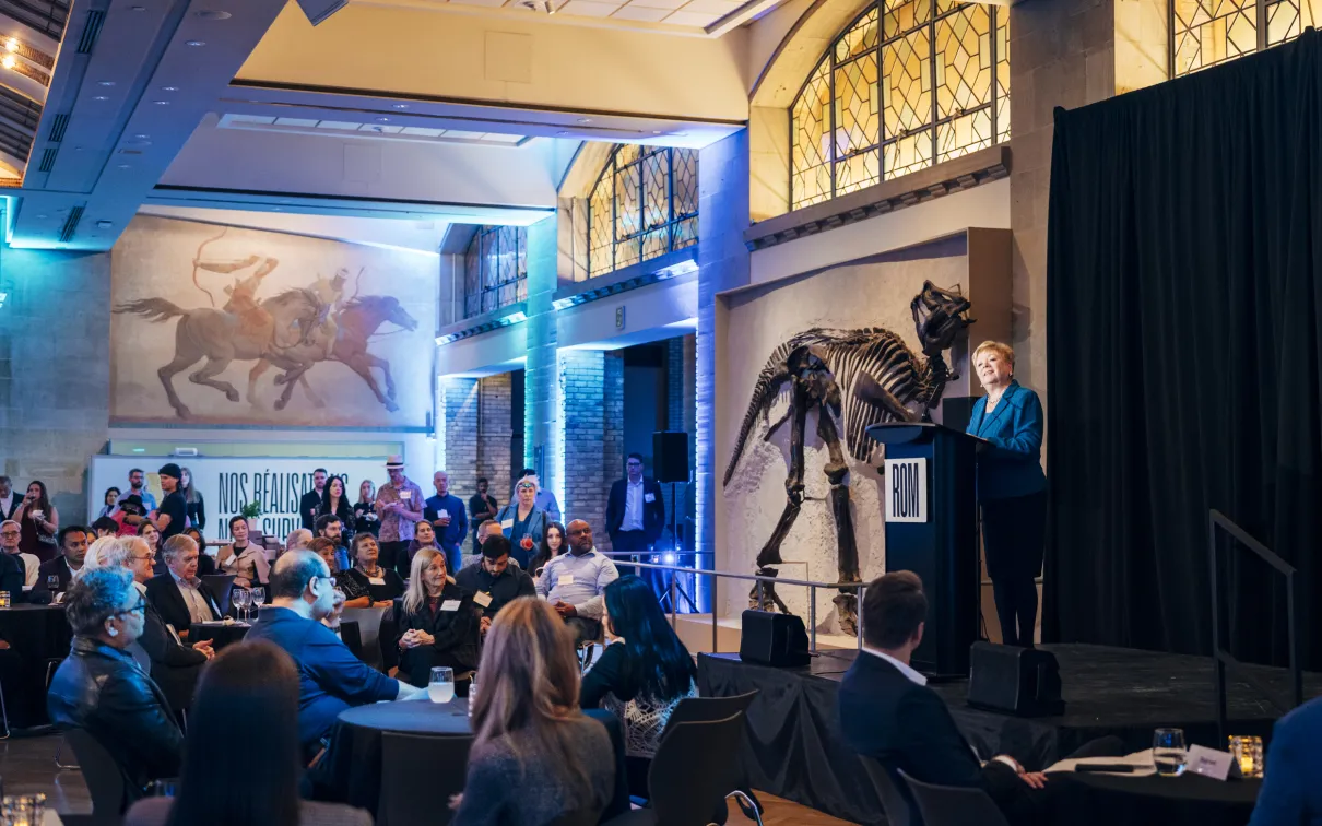 Audience seated in a large room listening to a speaker addressing the crowd from a stage with a large dinosaur skeleton.