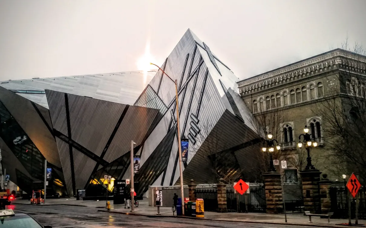 Modern angular glass and metal building facade of the Royal Ontario Museum with sunlight behind it, and a historic building in the background.