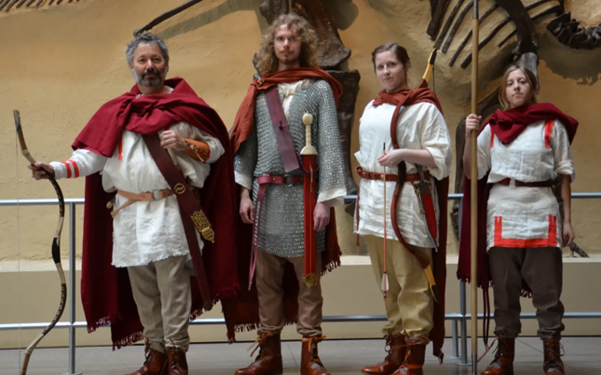 Four people dressed in ancient Roman soldier costumes standing indoors with a large dinosaur fossil mounted on a wall behind them.