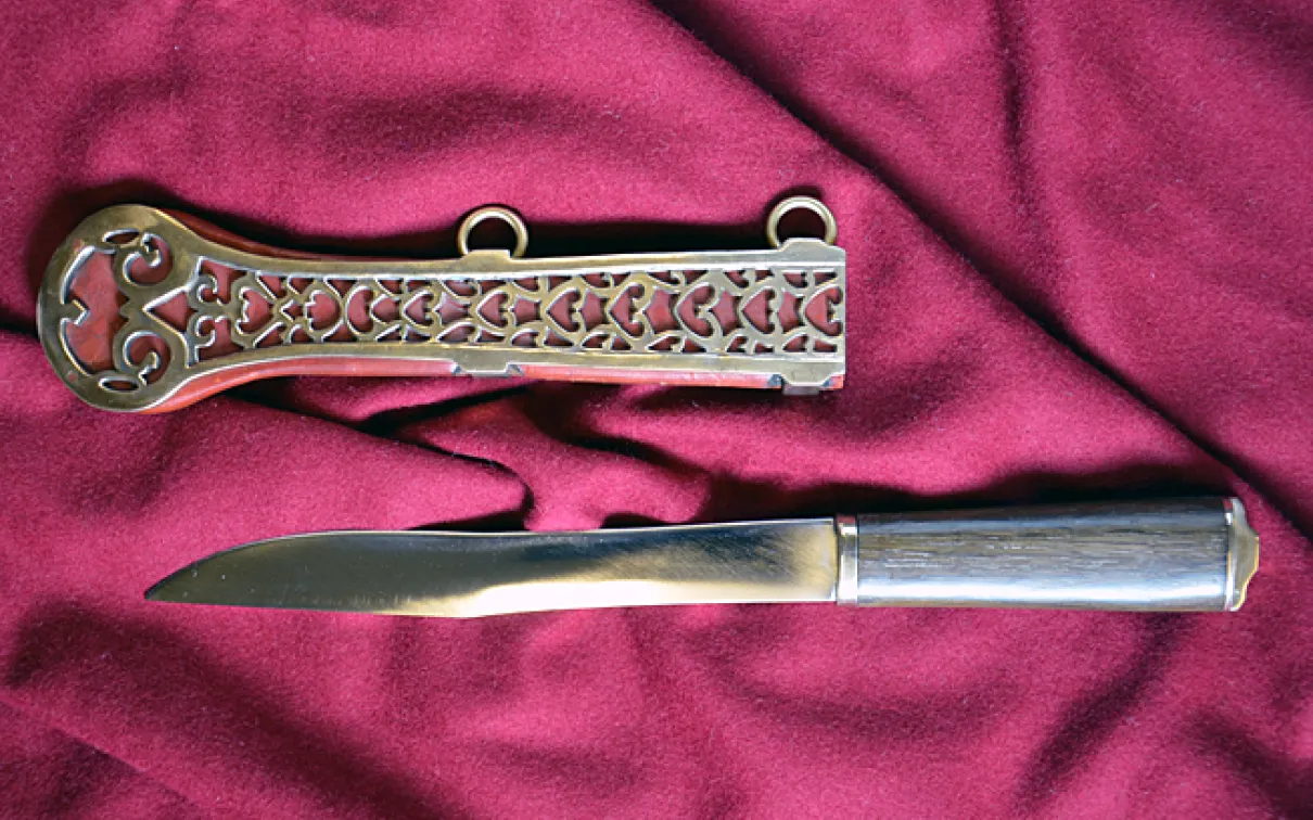 Steel knife with a straight blade and a polished metal handle next to an ornately decorated metal sheath on red cloth.