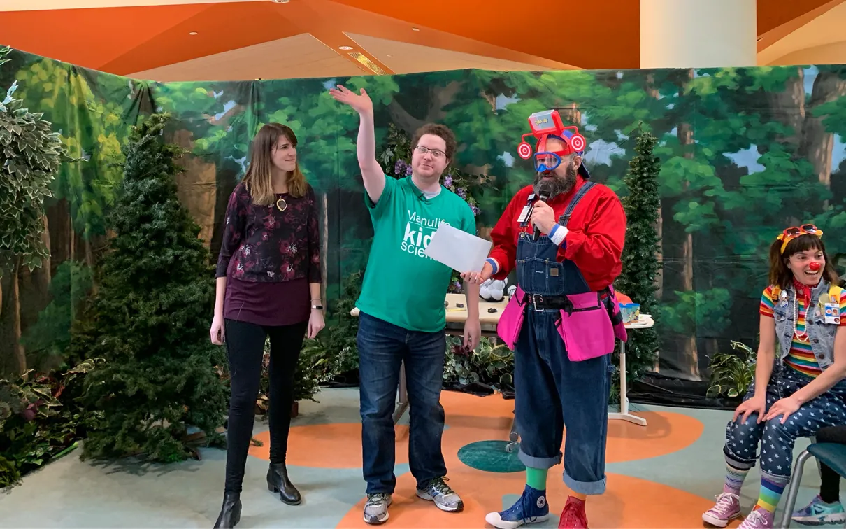 Three people stand on a colorful stage with a leafy green backdrop, one person in a red costume holds an item, and one person is seated on the right.