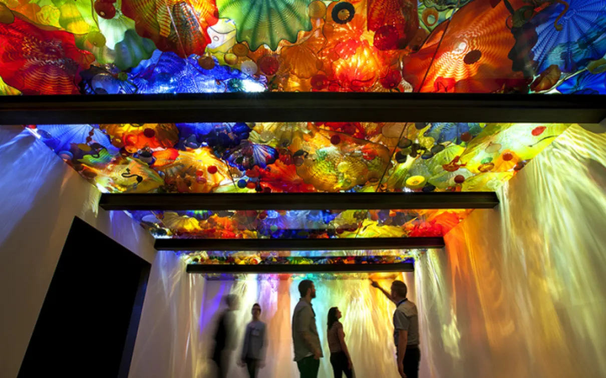 Visitors in the exhibition with glass sculptures displayed above a back lit glass ceiling.
