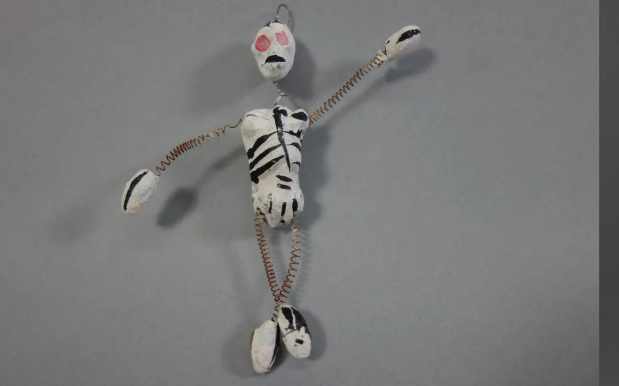 Small handcrafted figure with a white body painted with black markings, red mouth, and orange eyes, posed against a gray background.