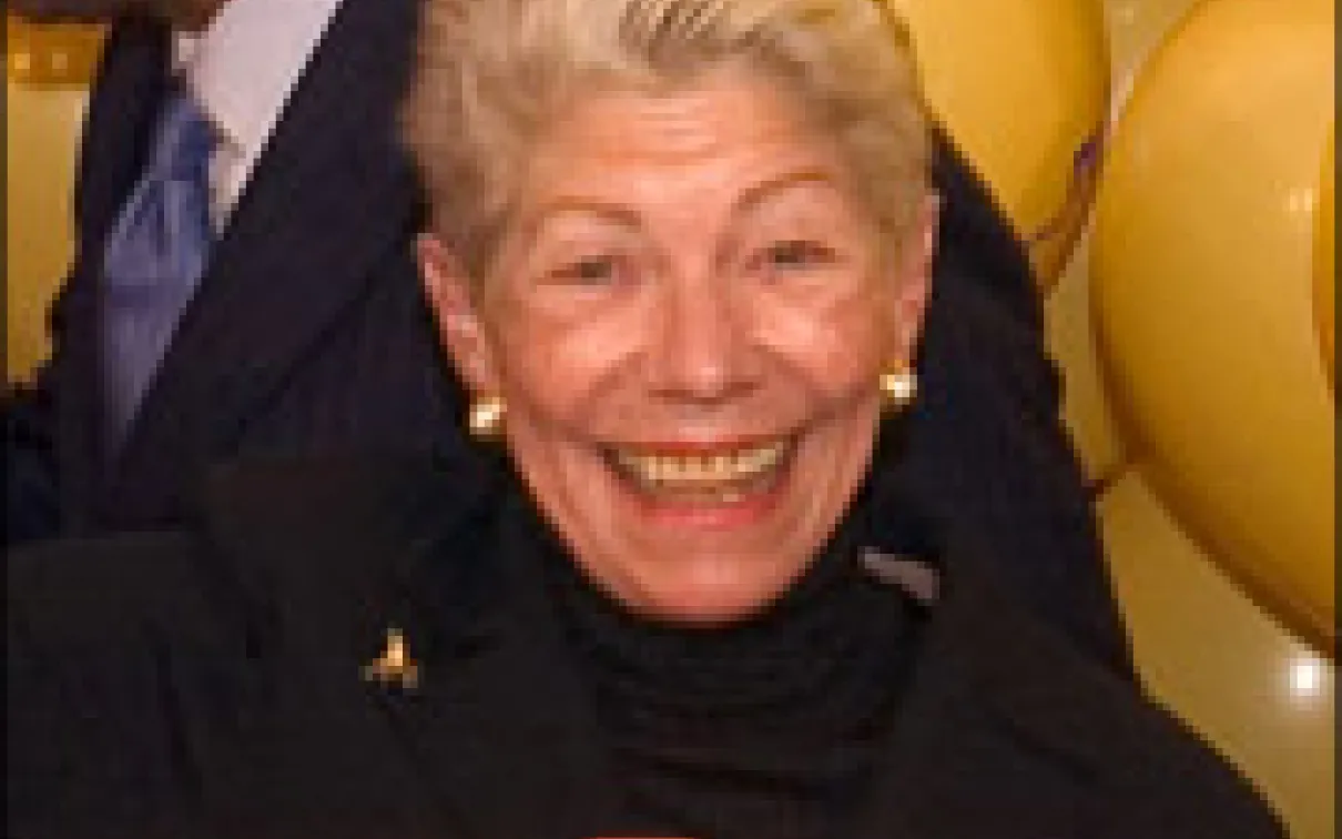 An elderly woman with short gray hair smiling, wearing a black top and pearl earrings.