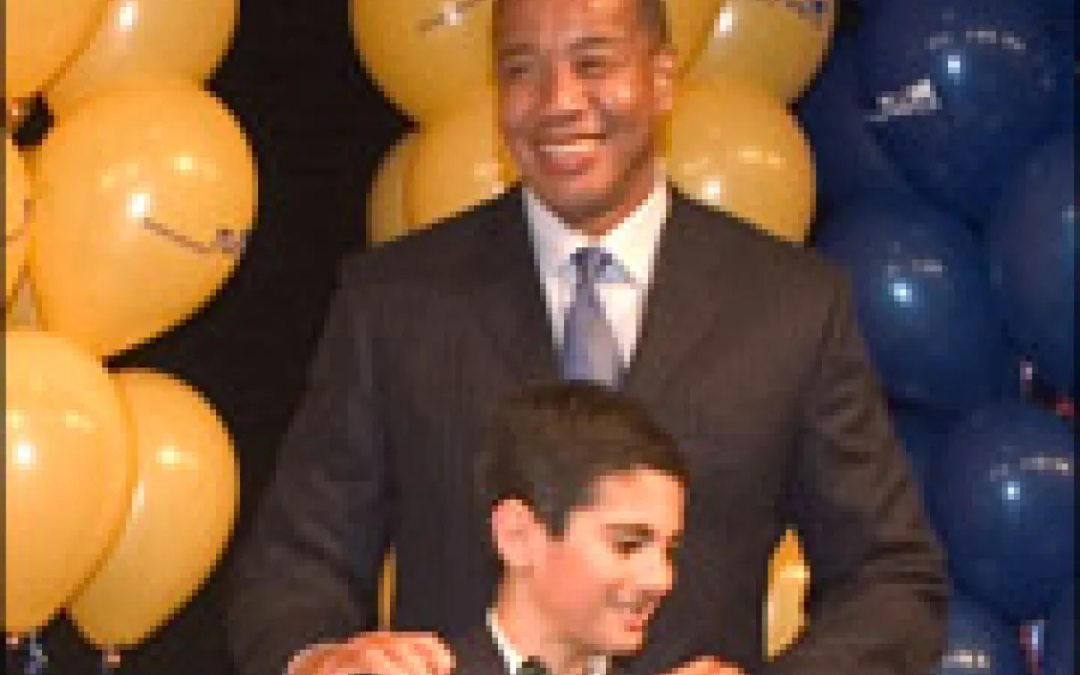 A man in a dark suit and tie stands behind a boy in a tuxedo with a bow tie, with yellow and blue balloons in the background.