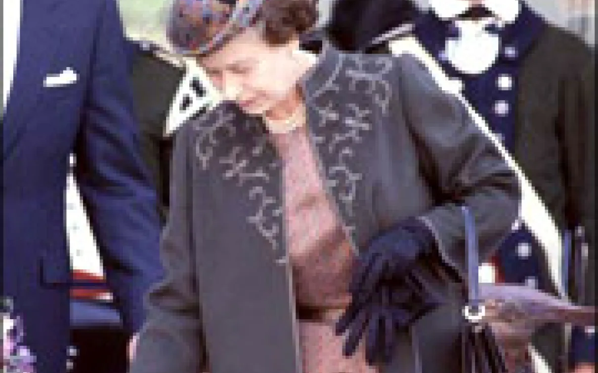 A woman in a dark coat with embroidery and matching hat holds a black handbag and wears black gloves.
