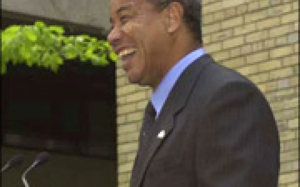 Man in a dark suit and tie smiling, standing in front of a yellow brick wall.