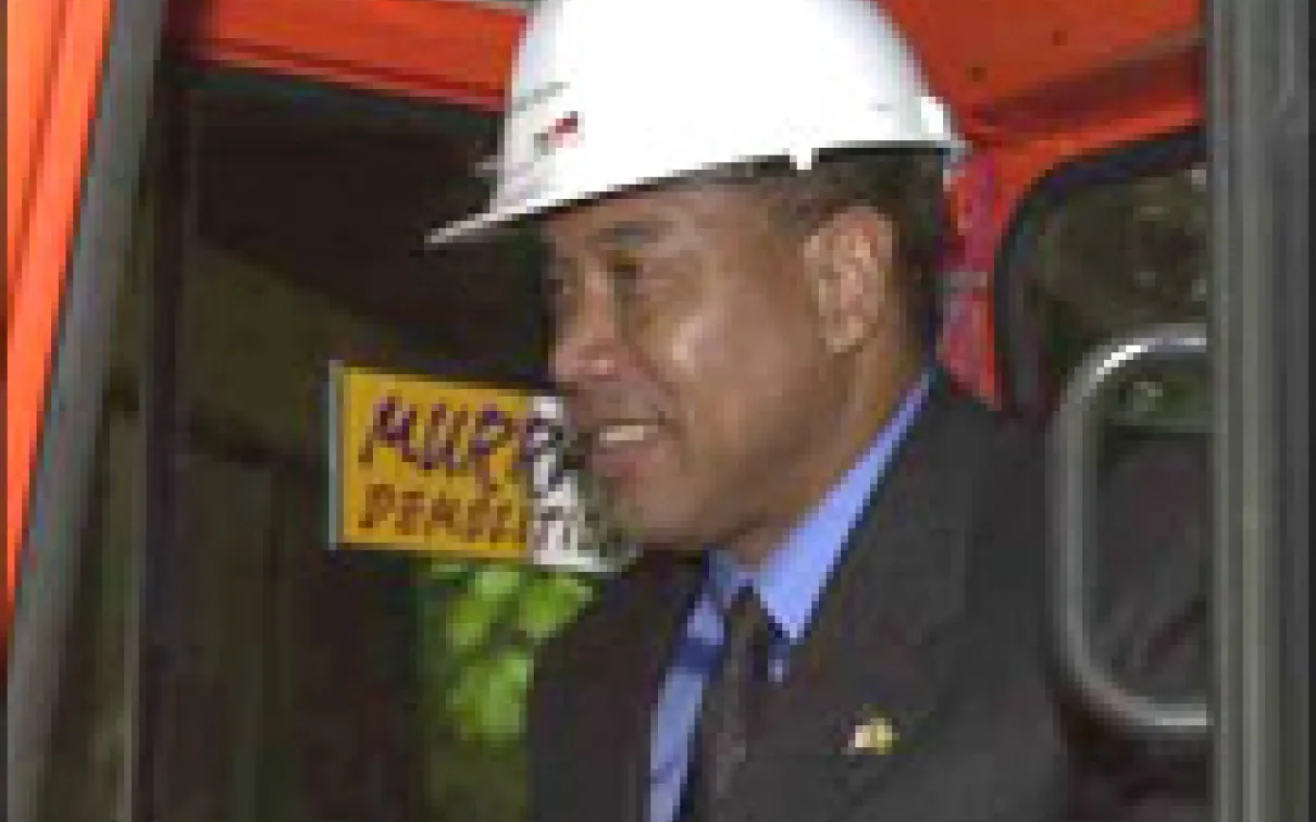 Man in a suit and tie wearing a white hard hat with a red logo and standing inside construction machinery.