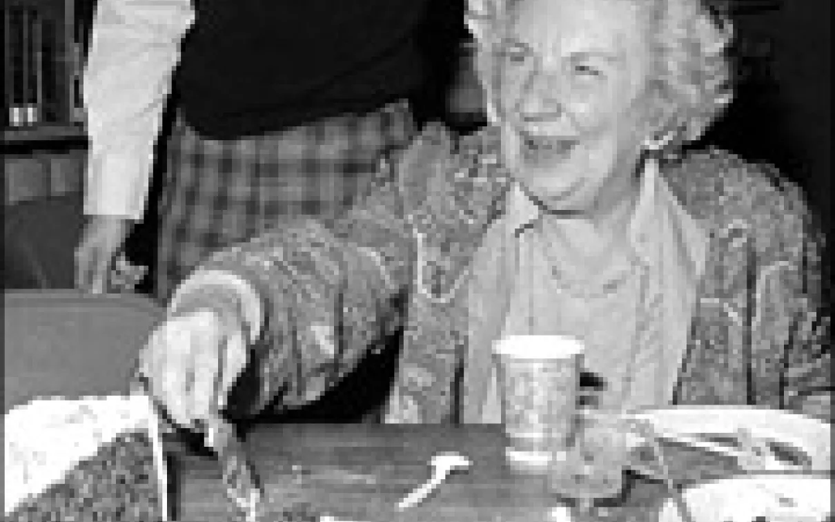An elderly woman sitting at a table, smiling and cutting a cake with a knife.