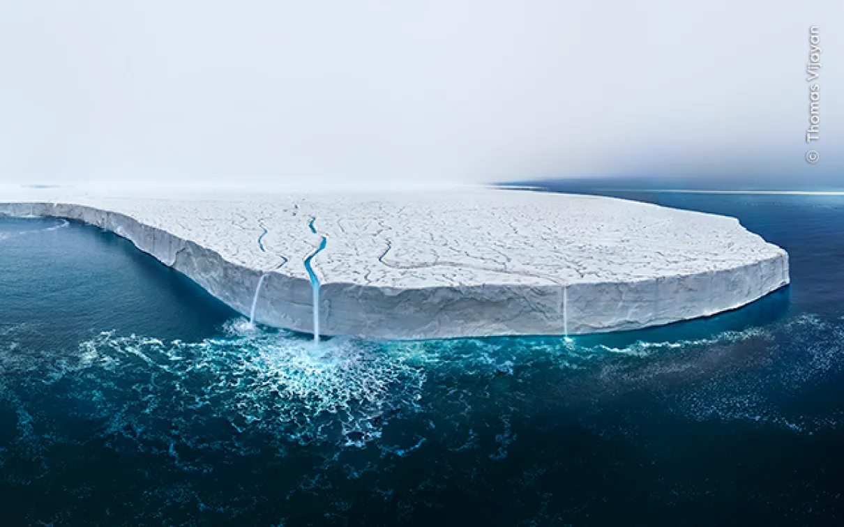 A large ice shelf floating on calm, dark blue ocean water under a clear sky.