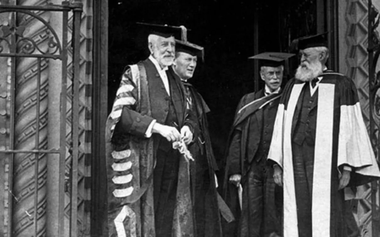 Four men dressed in academic regalia standing on a building entrance decorated with wrought iron gates.
