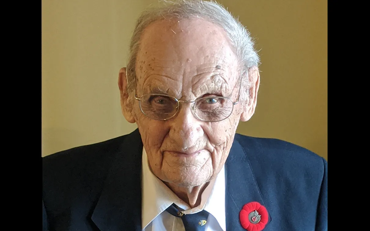 Elderly man wearing a dark blazer, striped shirt, and tie with a red poppy pin on his lapel.