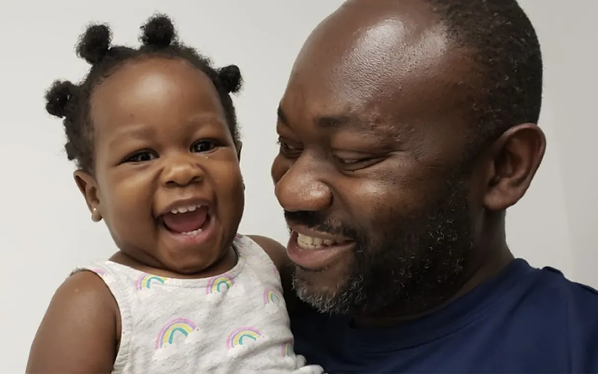 A smiling man in a blue shirt holding a laughing toddler wearing a white tank top with rainbow patterns.
