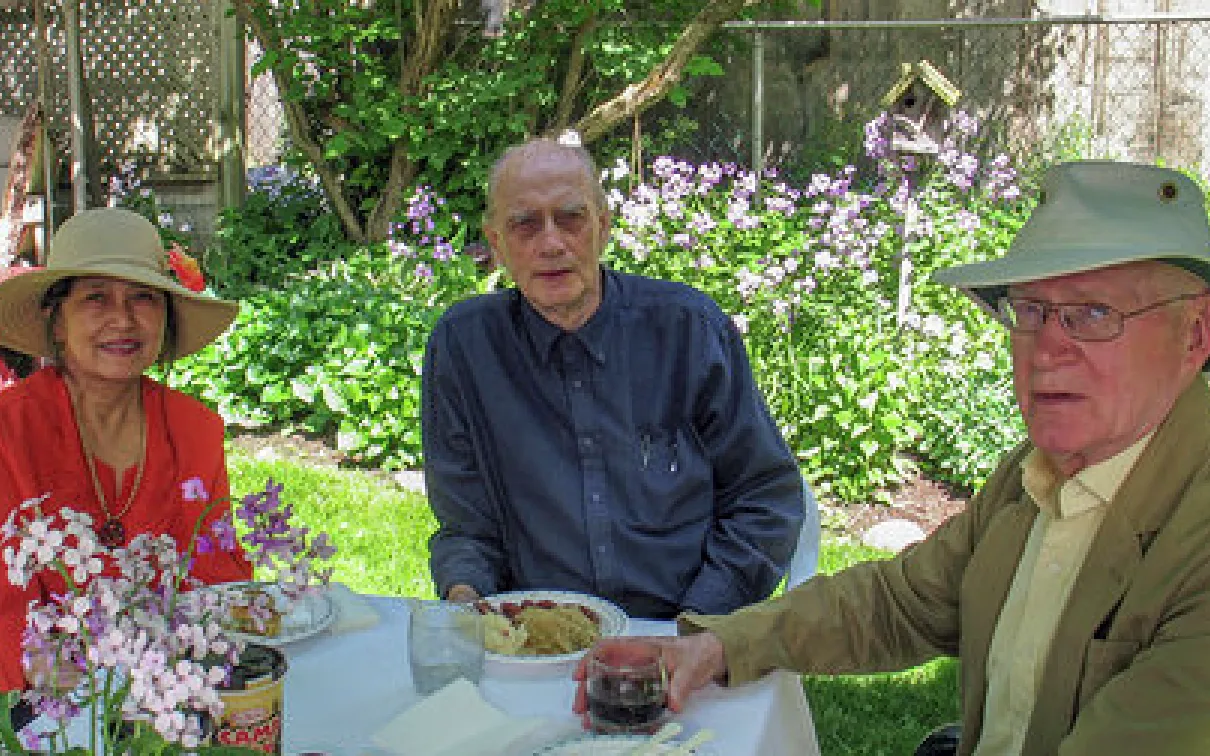 Three older adults sitting outdoors at a table with flowers and greenery in the background.