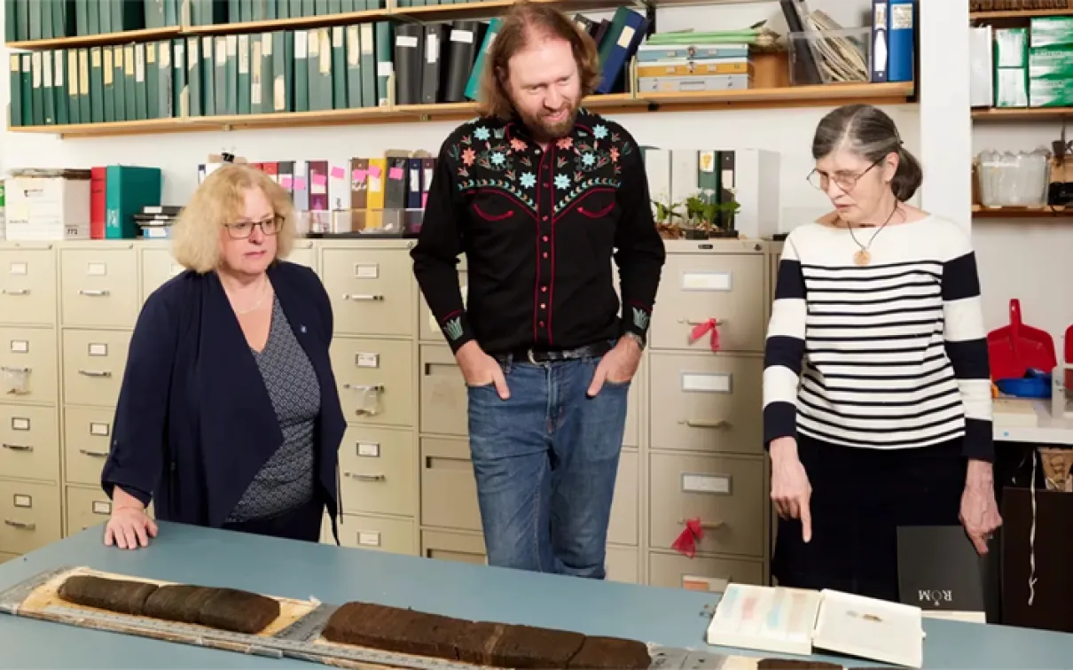 Three people standing behind a table with artifacts and documents in a room filled with shelves and filing cabinets.