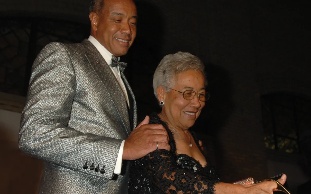 A man in a silver suit jacket stands behind a woman in glasses and a black lace dress.