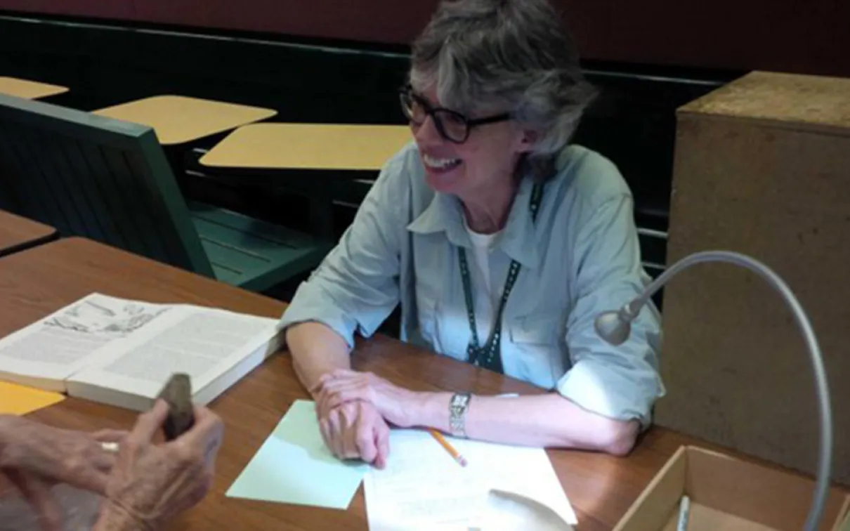 A woman sitting at a desk writes on paper with open books and a black pen visible on the table.
