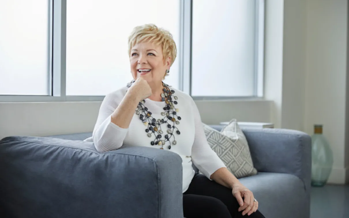 Portrait of Janice Price smiling and seated on a gray sofa, wearing a white blouse and a statement necklace.