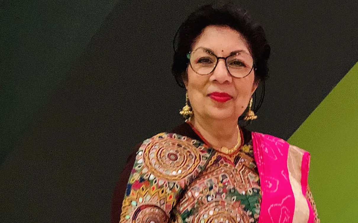 A woman wearing a brown patterned top, gold necklace, glasses, and a bright pink shawl on her left shoulder against a green and black background.