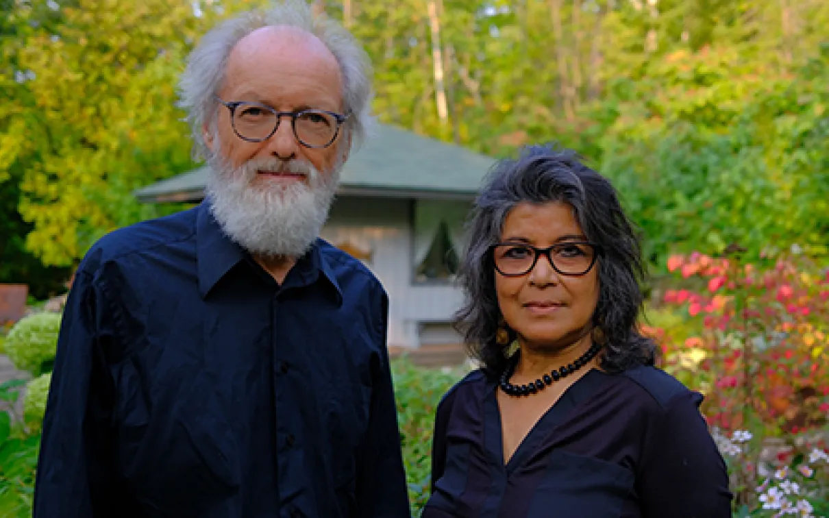 Portrait of an older man with white hair and beard wearing glasses and a black shirt, and a middle-aged woman with dark hair and glasses wearing a black top, standing outdoors with green trees and a pavilion in the background.