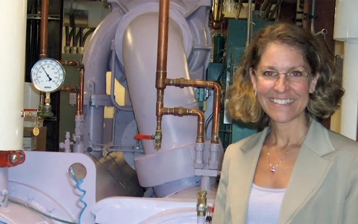A woman with glasses and curly hair stands smiling in front of industrial pipes and gauges.