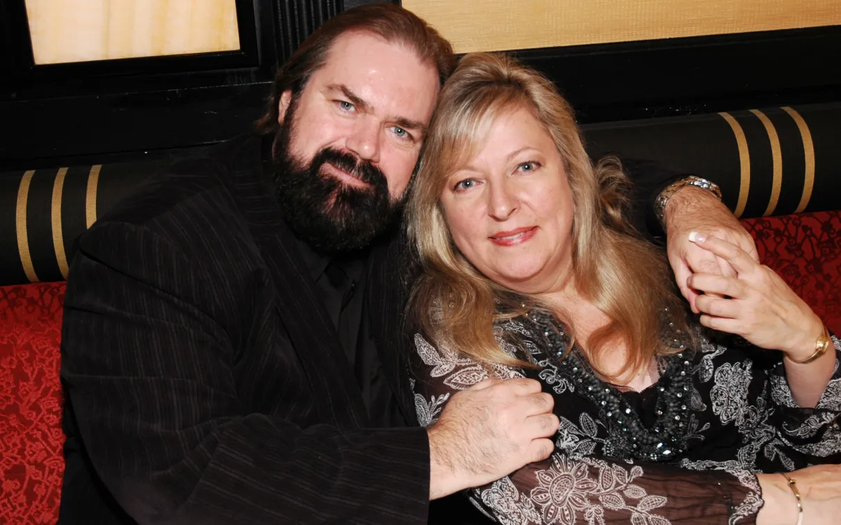 A man with a beard and a woman with blonde hair sit together on a striped couch, both looking at the camera.