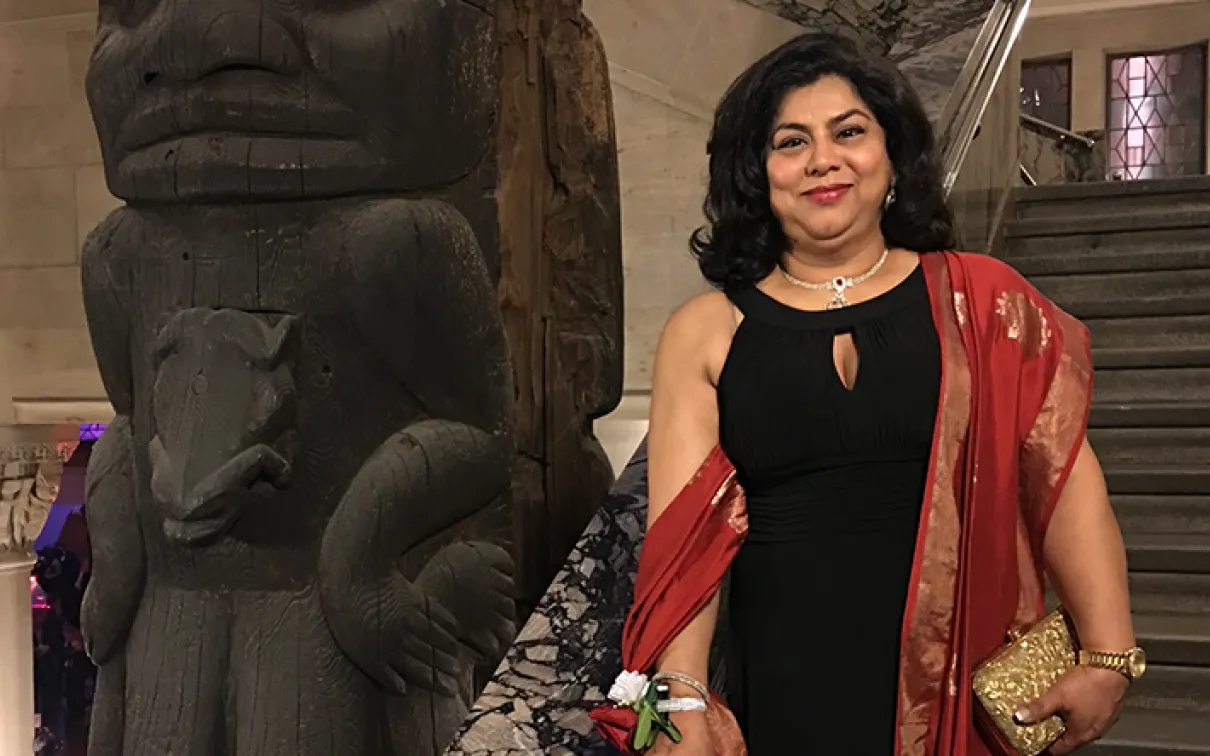 A woman in a black dress with a red shawl stands beside a large carved wooden totem pole inside a building.