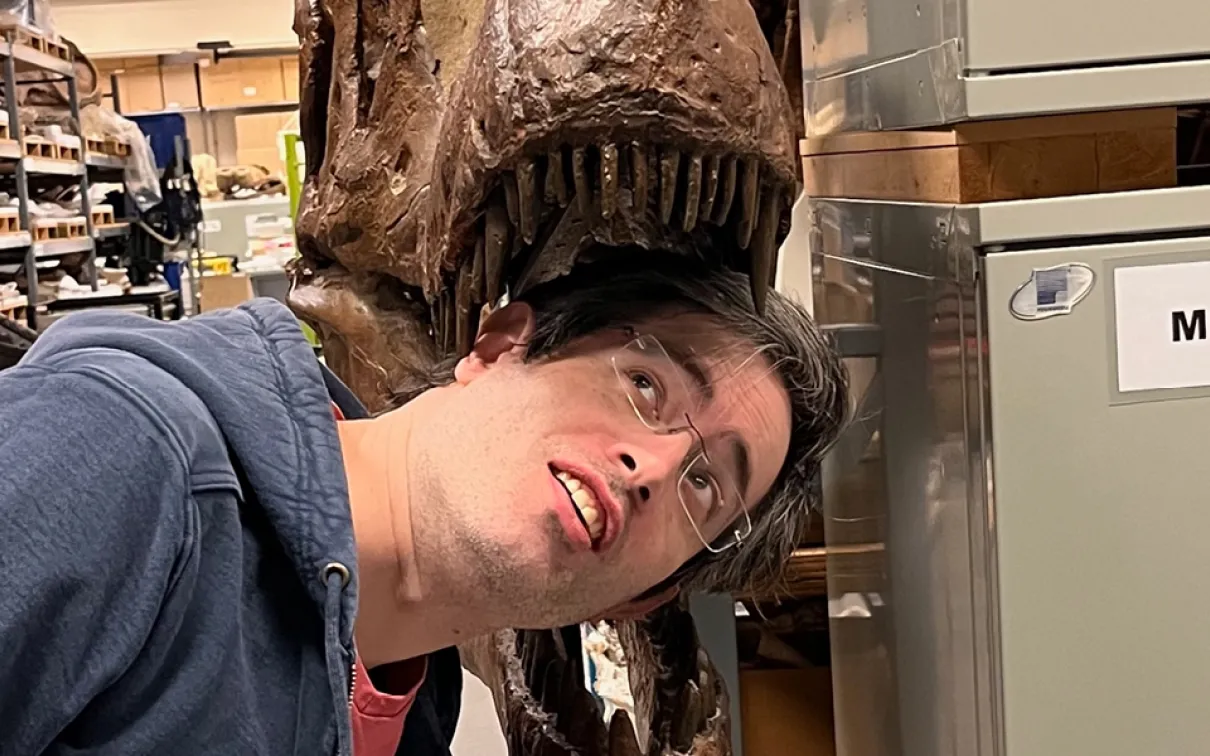 A person with dark hair leans their head toward the jaws of a large dinosaur skull mounted on a display stand.