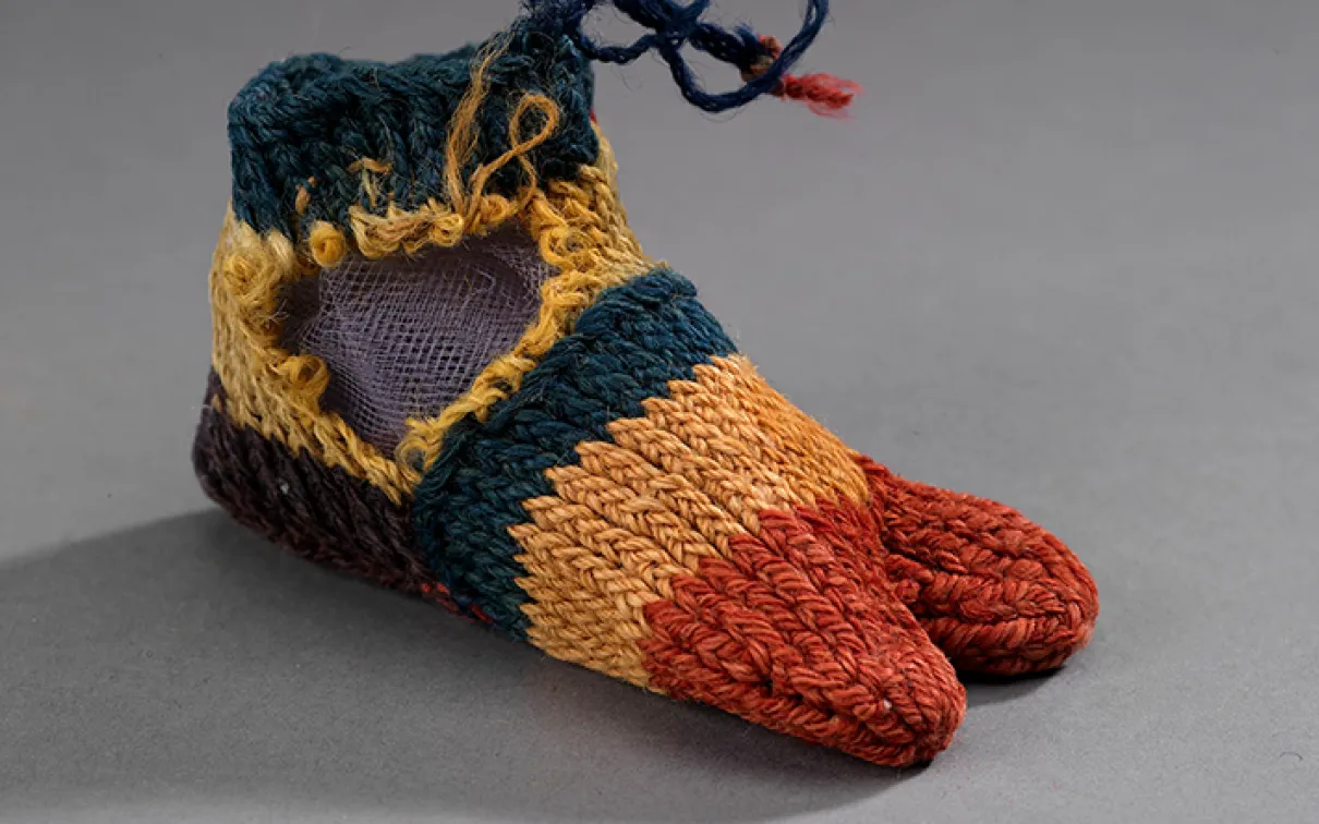 Woven baby shoes with orange toes, yellow and green bands, and blue laces on gray background.