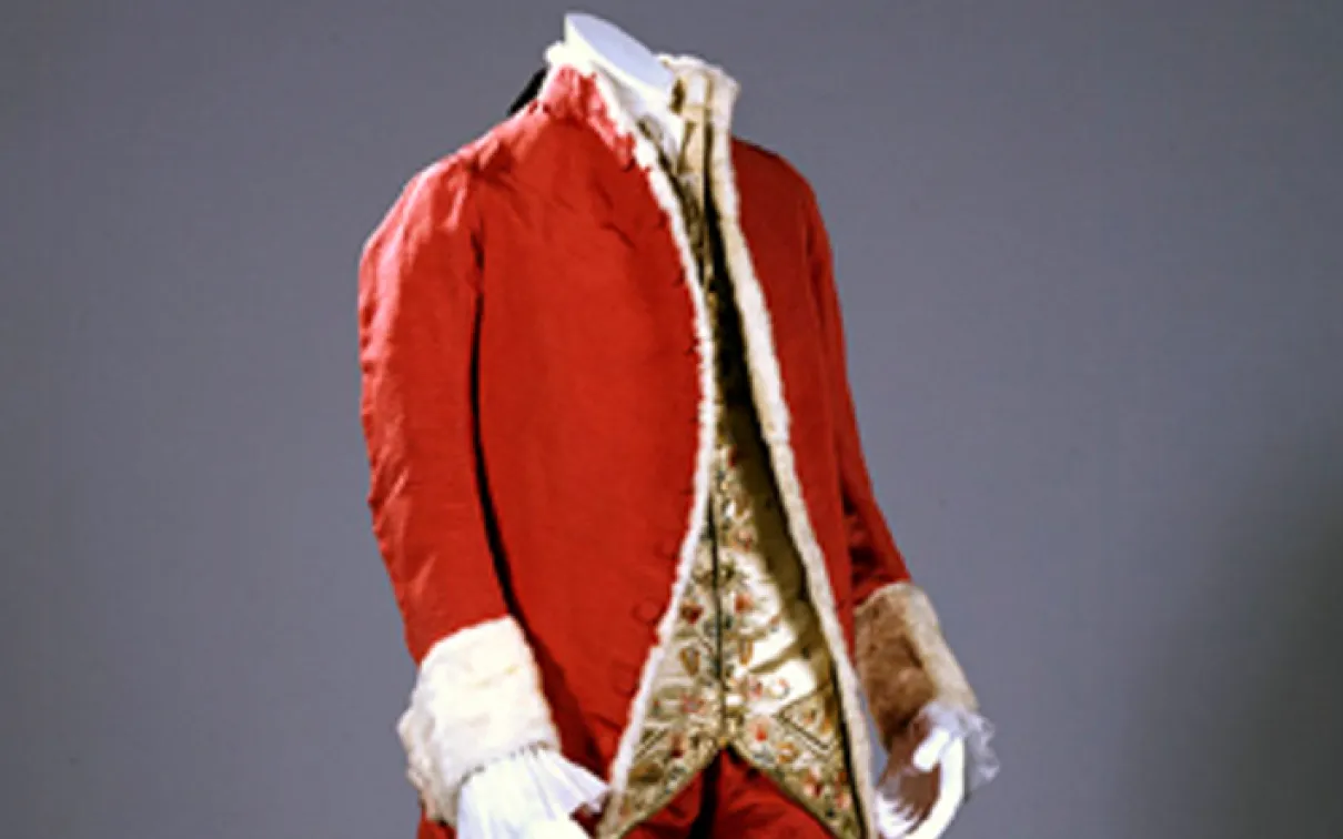 Red 18th-century coat with white lace cuffs and a patterned waistcoat underneath.