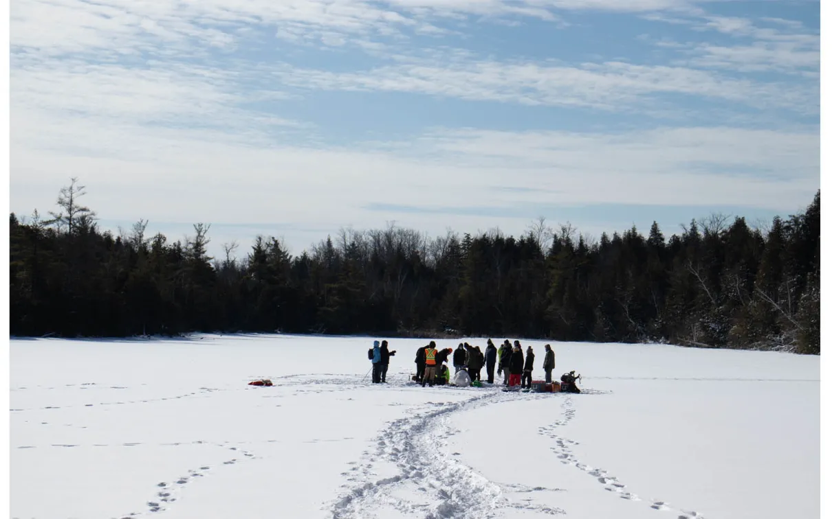 Group of people gathered on a snowy frozen lake surrounded by trees under a partly cloudy sky.
