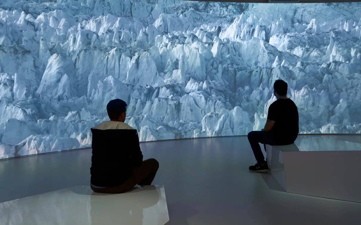 Two people sit on benches facing a large digital wall displaying a blue-hued glacier scene.