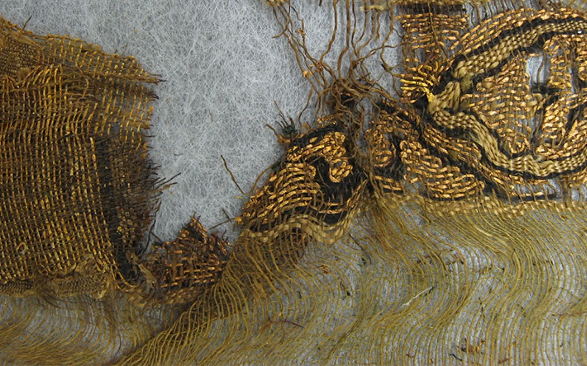 Fragment of ancient textile showing loose threads and woven fabric in brown and beige tones.