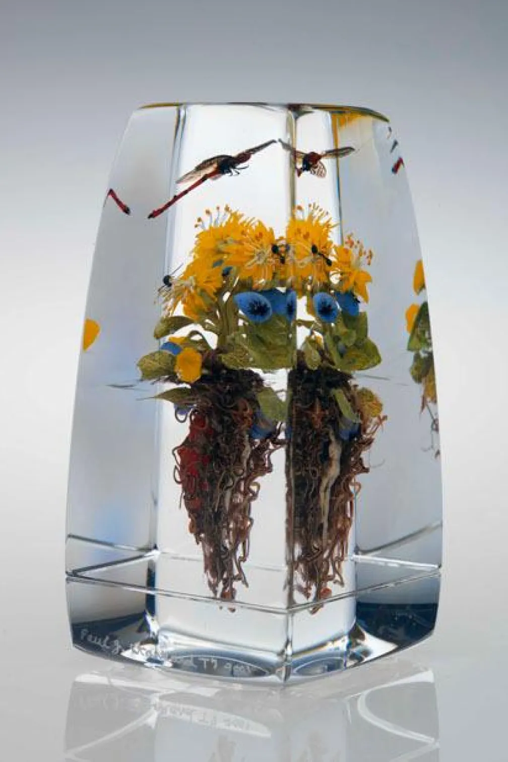 Yellow flowers with green leaves and brown roots encased in a clear rectangular resin block.