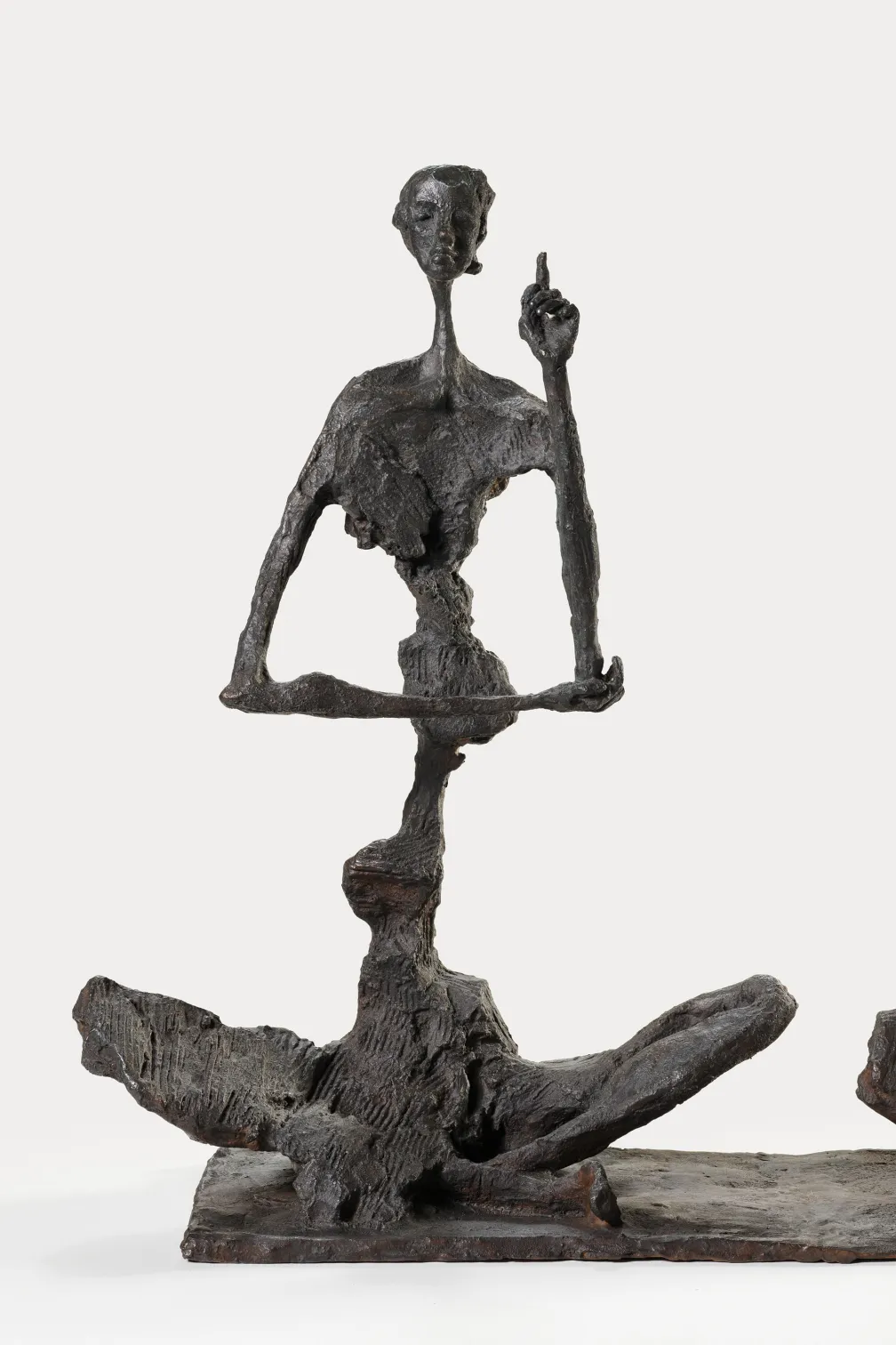 Bronze sculpture of two elongated, abstract figures stacked vertically, with the upper figure seated cross-legged and raising one hand.