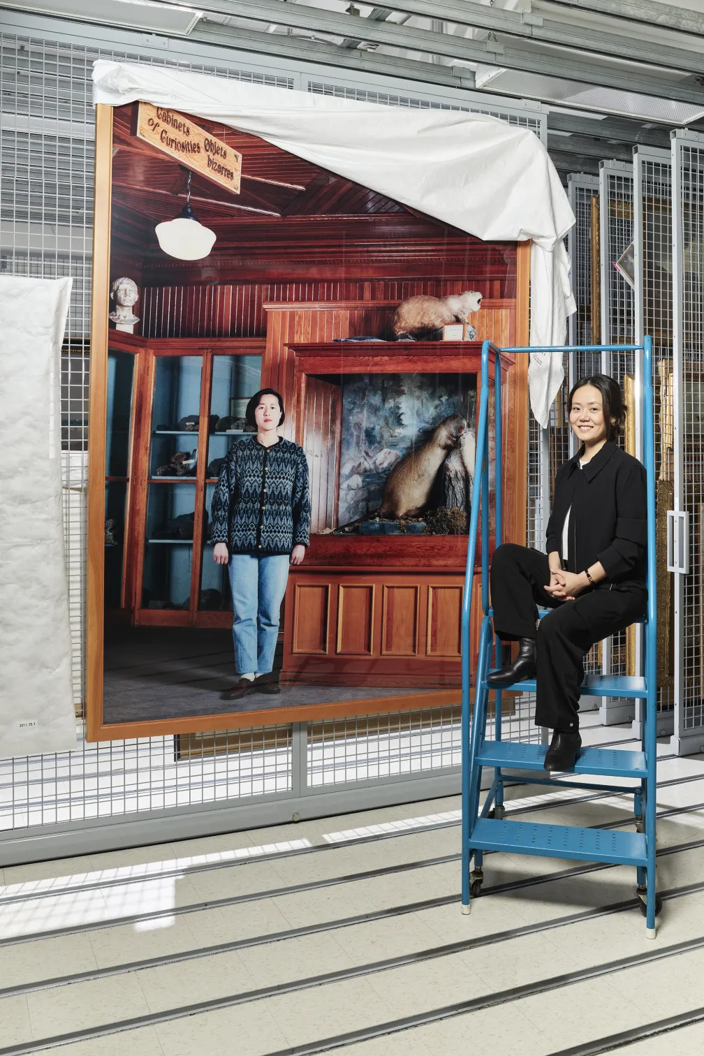 Two women pose near a large photographic print hanging from a metal grid; one woman stands in front while the other sits on a ladder.
