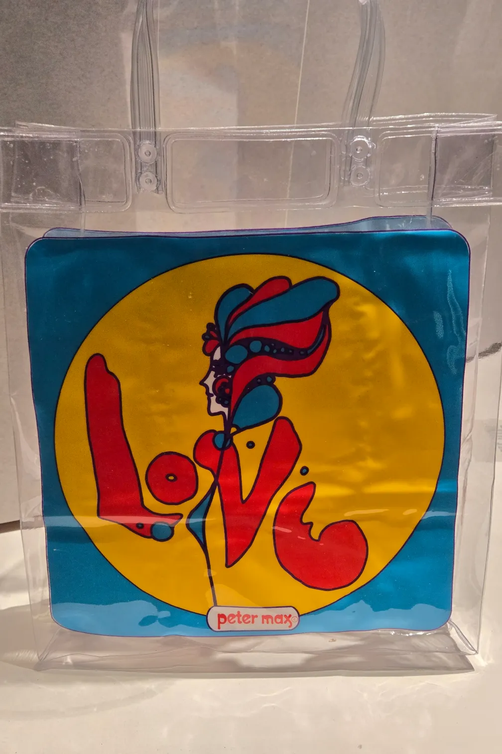 Un sac en plastique transparent dans un coffret de musée. Sur le devant du sac figure l'image d'une femme dans un cercle jaune sur fond bleu. Les armes et le manteau de la femme épellent également le mot AMOUR. Peter max est imprimé au bas du sac.