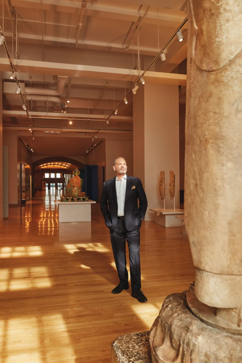 A man in a black tuxedo standing in a spacious gallery with wooden floors, stone columns, and display cases.