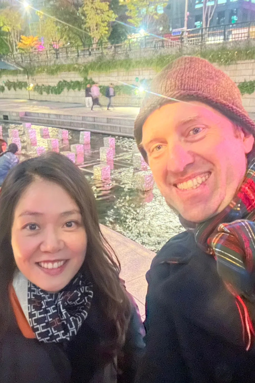 Two people smiling outdoors at night with festive string lights and reflections on water behind them.
