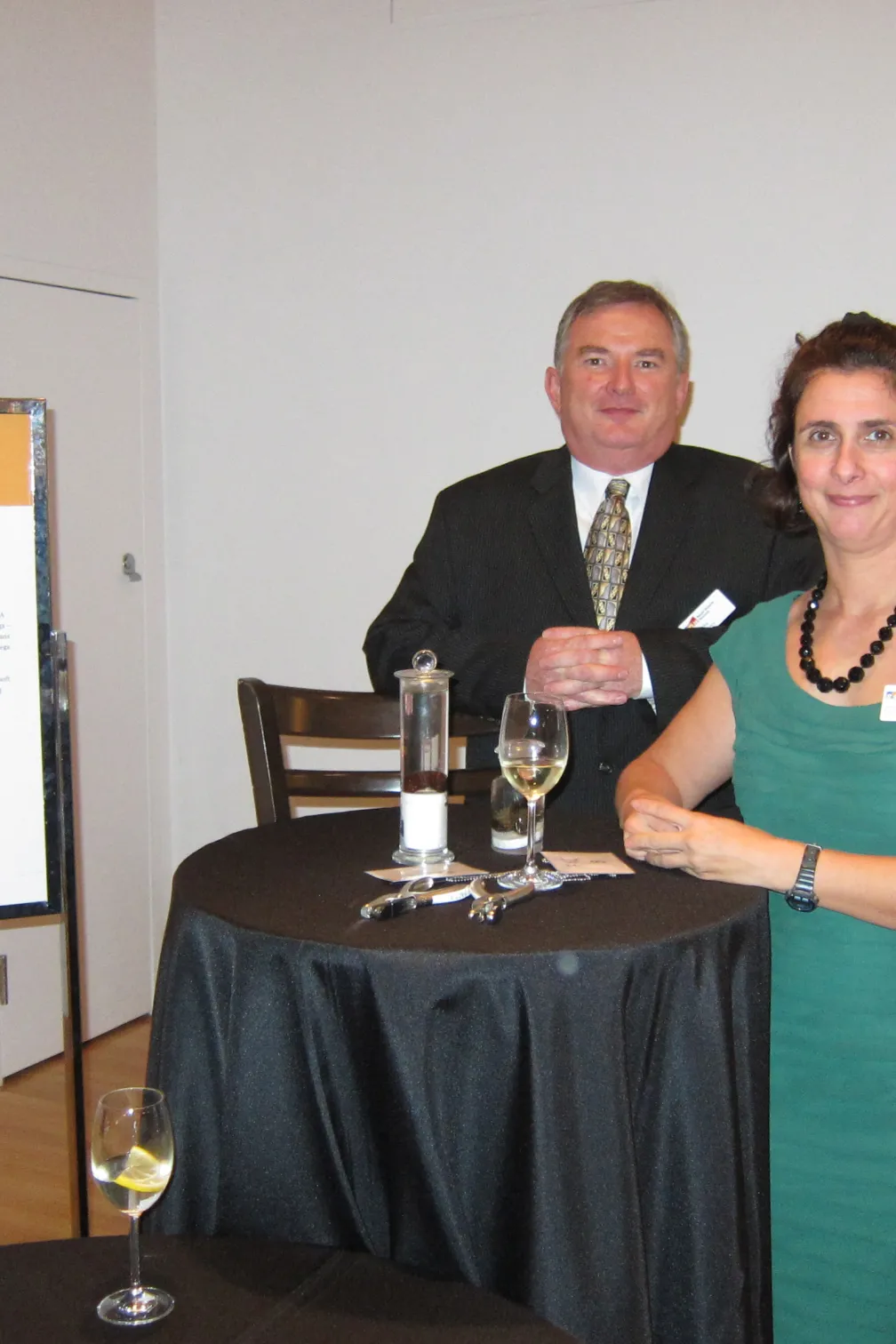 Two people stand next to a high round table with glasses and drinks in a room with a white wall and a sign labeled "No. 1."