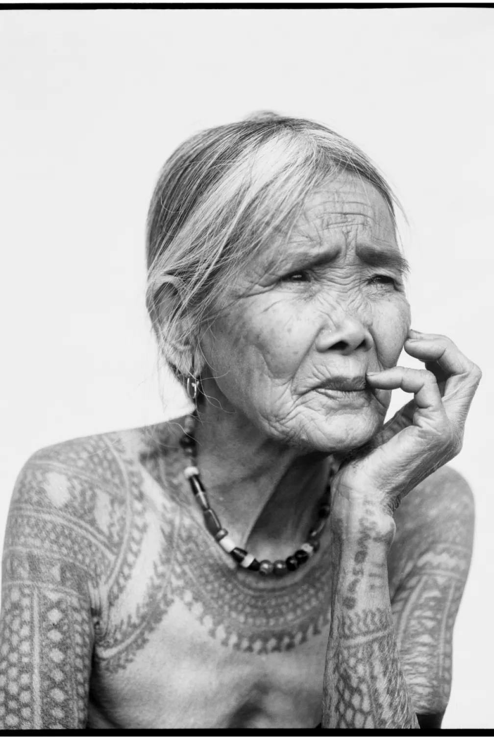 Elderly woman with traditional tattoos on her arms and upper chest, resting her chin on her hand.
