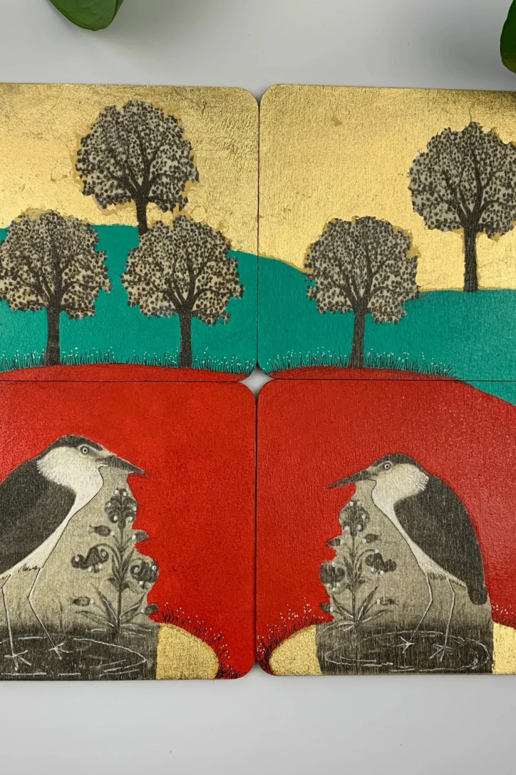 Four square coasters with textured art: the top two show black trees on yellow and green backgrounds; the bottom two show black and white birds on red and yellow backgrounds.