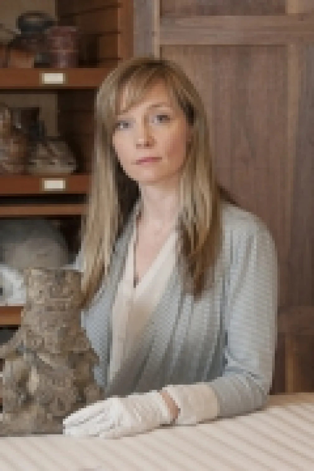 A woman wearing white gloves poses with an ancient carved stone artifact on a table, with wooden shelves holding pottery in the background.